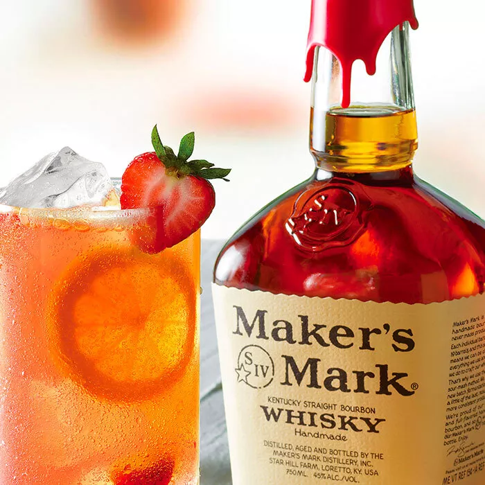 A tall glass contains a Kentucky Buck cocktail and is garnished with strawberries, lemon pinwheels, and a mint sprig. A bottle of Maker's Mark is next to the cocktail on the right.