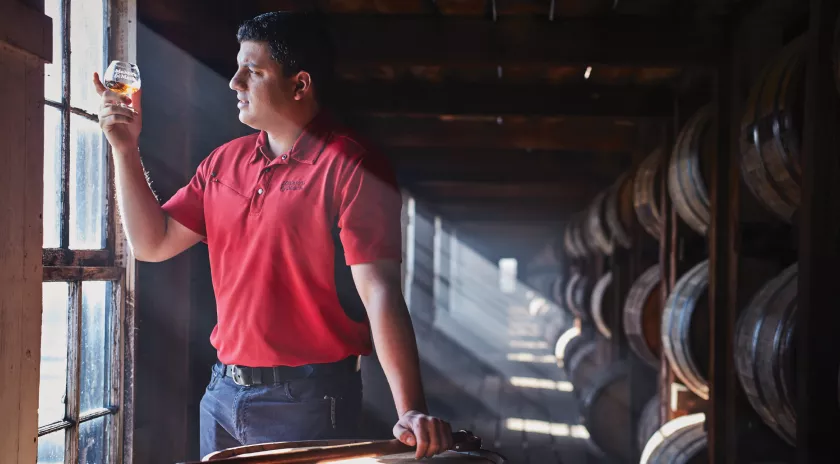 Man staring out the window holding glass of Makers Mark in distillery with barrels behind him in an early morning sun