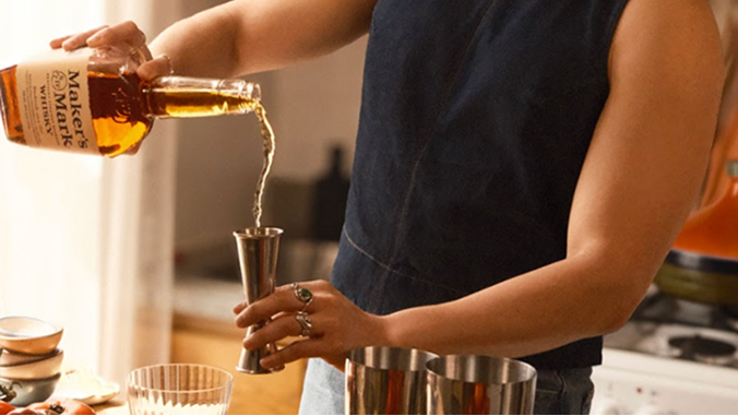 a person in a sleeveless dark shirt pours Maker's Mark whiskey from a bottle into a glass while standing in a bright kitchen.
