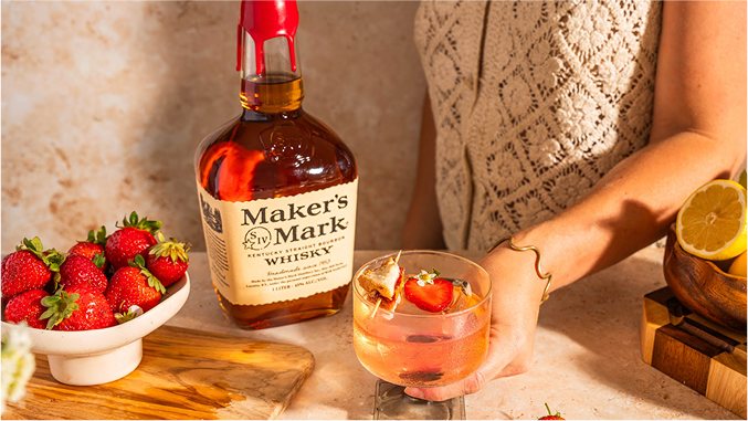 A hand holds a cocktail garnished with strawberries, beside a bottle of Maker's Mark whisky and a bowl of fresh strawberries.