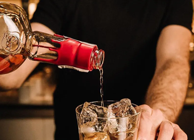 A bartender pours amber liquid from a bottle into a glass filled with ice, set against a softly lit bar background.
