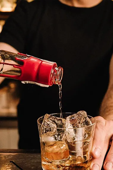 A bartender pours amber liquid from a bottle into a glass filled with ice, set against a softly lit bar background.