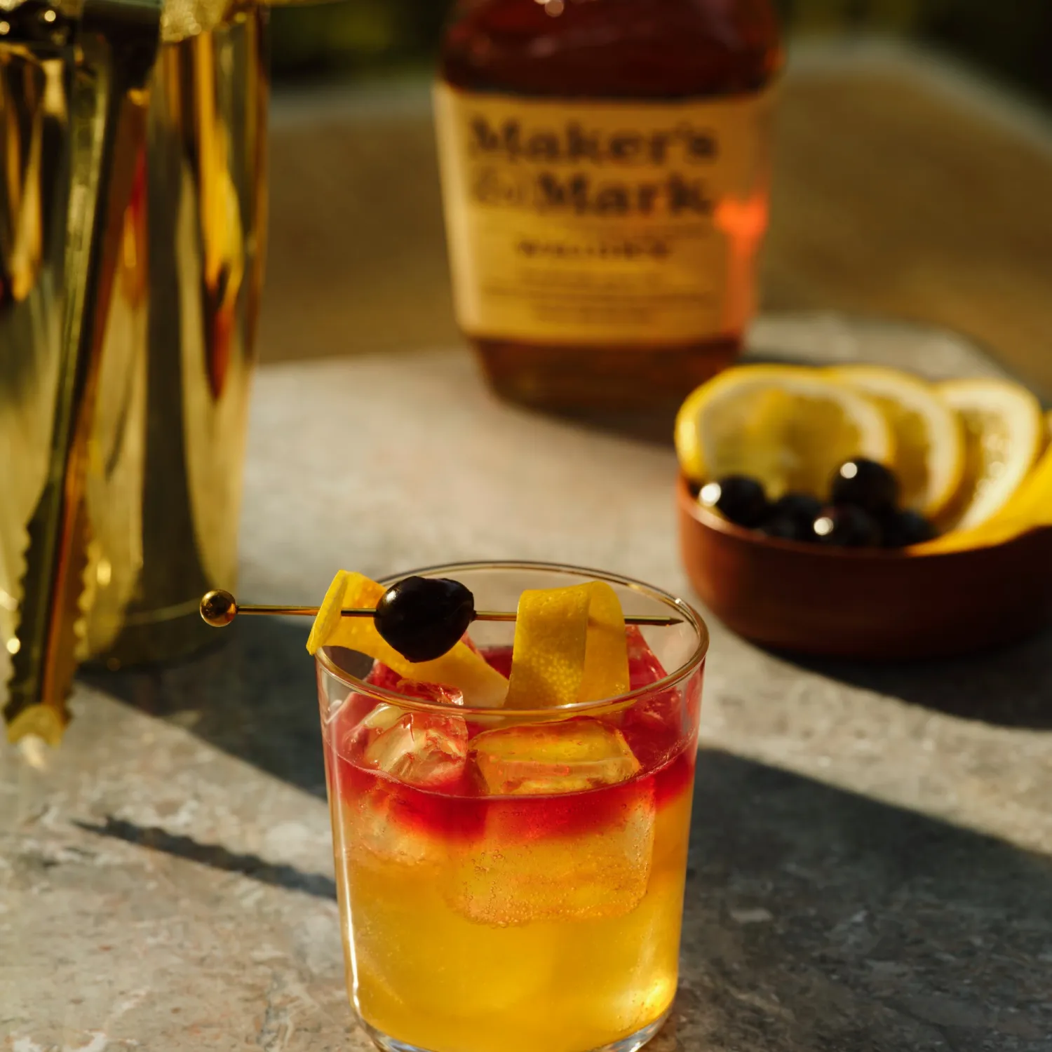 A glass of whiskey cocktail with ice, garnished with a lemon peel and cherry, sits on a table. A whiskey bottle, bowl of lemon slices, and a golden shaker are in the background. The setting conveys a warm, elegant mood.