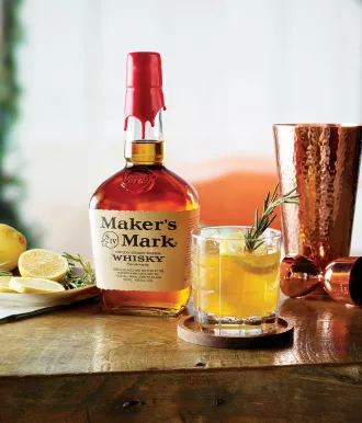 Rosemary maple sour cocktail with garnish on table next to a bottle of makers mark