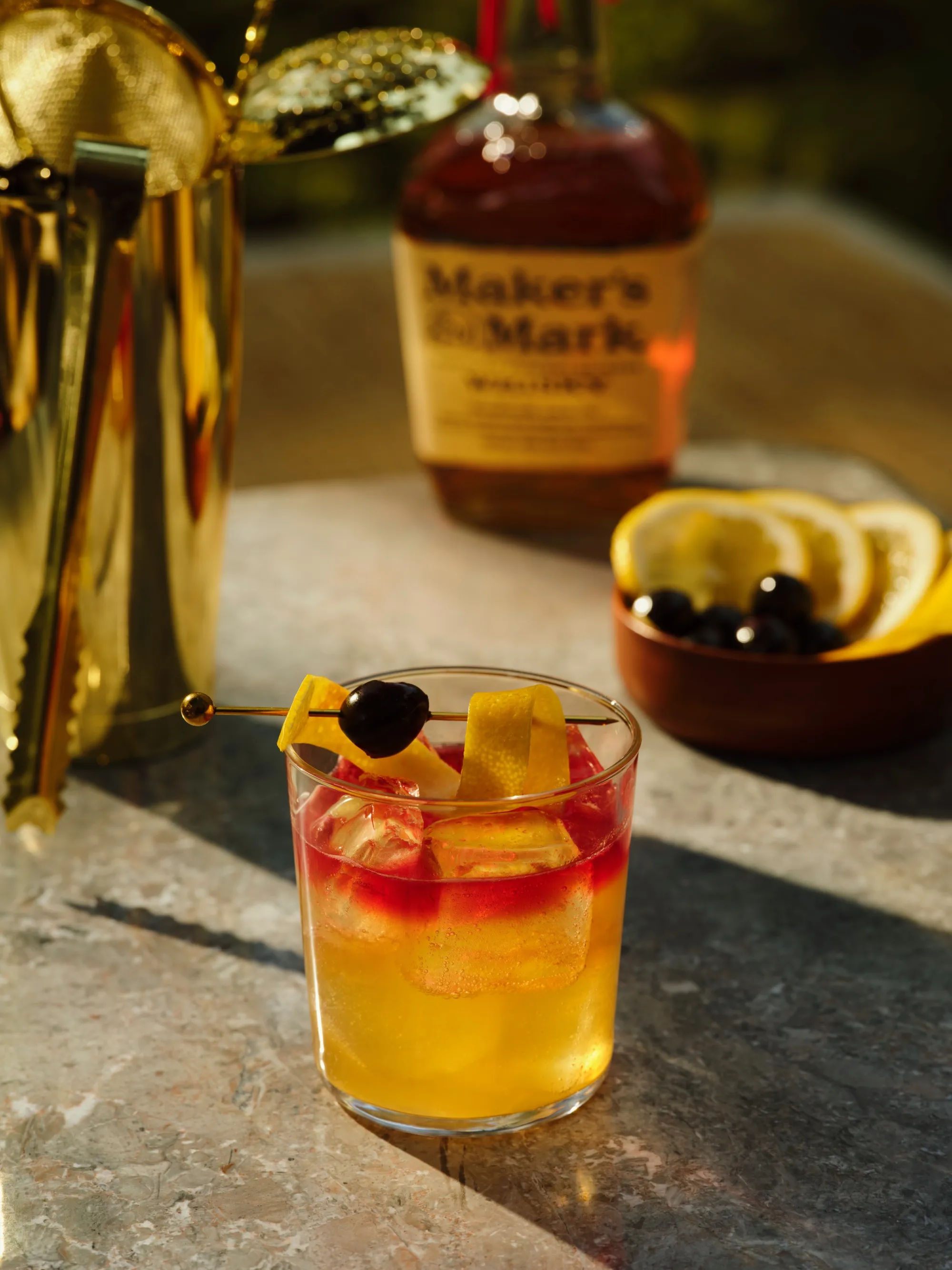 A glass of whiskey cocktail with ice, garnished with a lemon peel and cherry, sits on a table. A whiskey bottle, bowl of lemon slices, and a golden shaker are in the background. The setting conveys a warm, elegant mood.