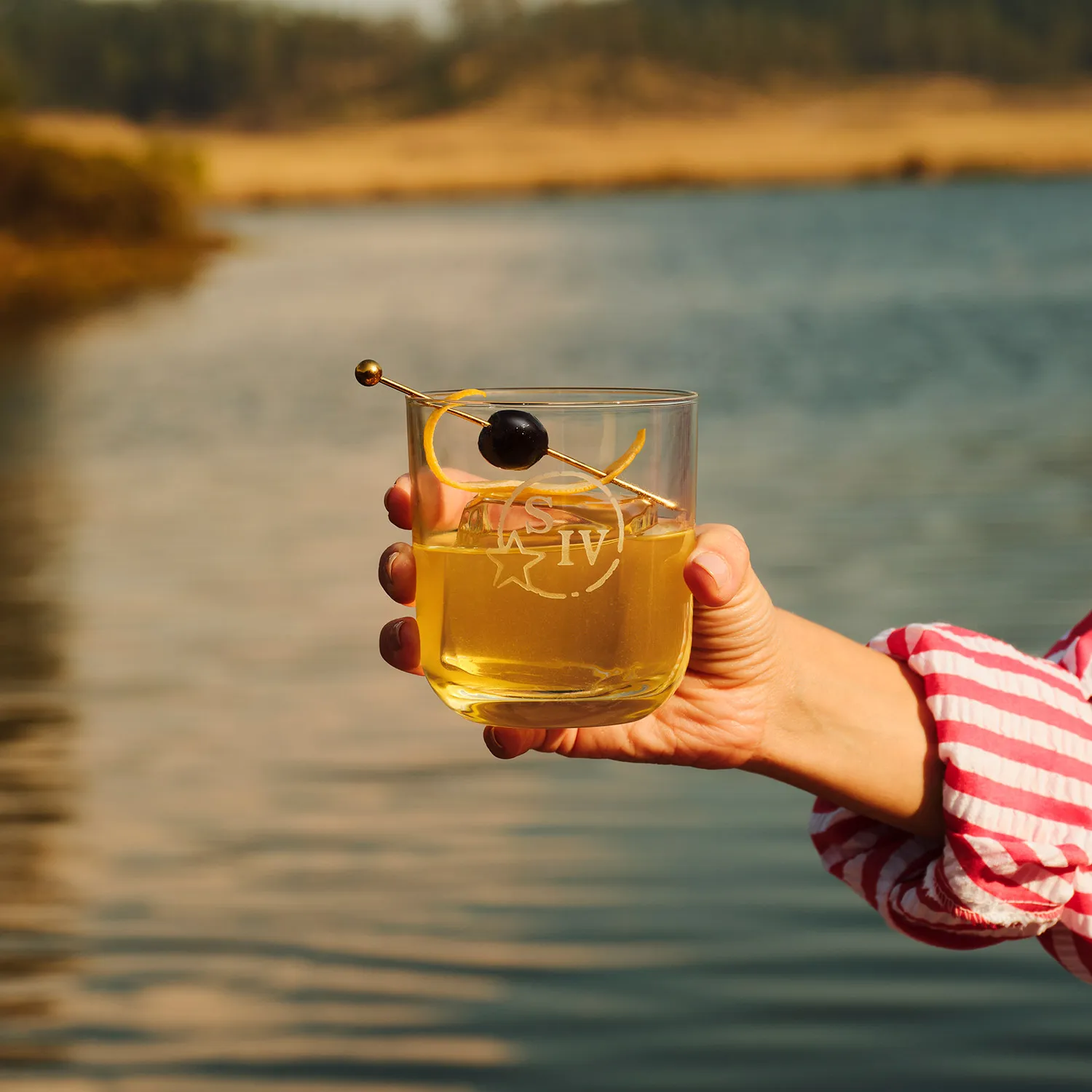 A hand holds a Bourbon Sour cocktail with a cherry and orange peel garnish by a lake, Cocktail Whisky Maker's Mark.