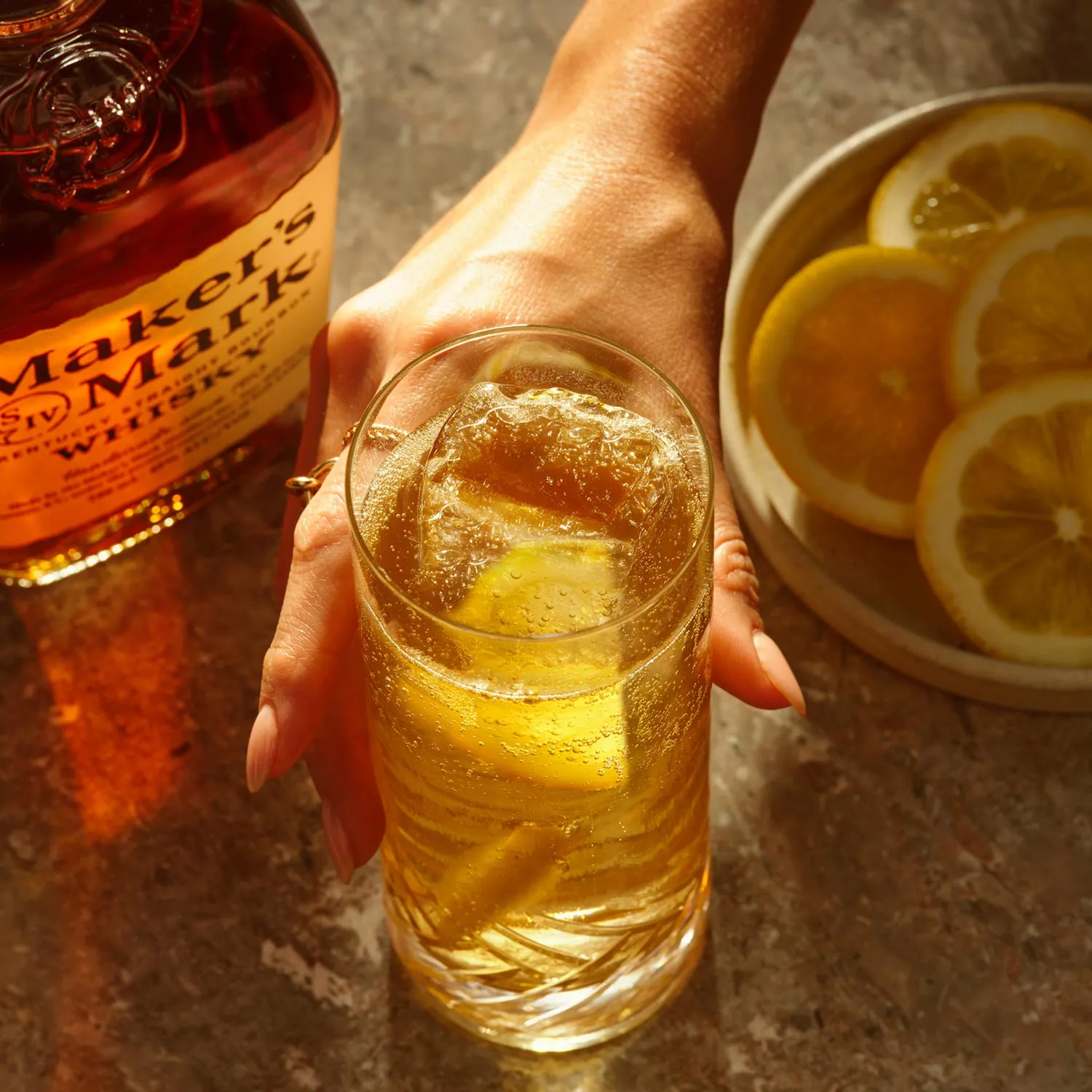 A hand reaches for a Sour Highball cocktail with a lemon slice and ice, near a bottle of Whisky Maker's Mark, Cocktail Whisky Maker's Mark.