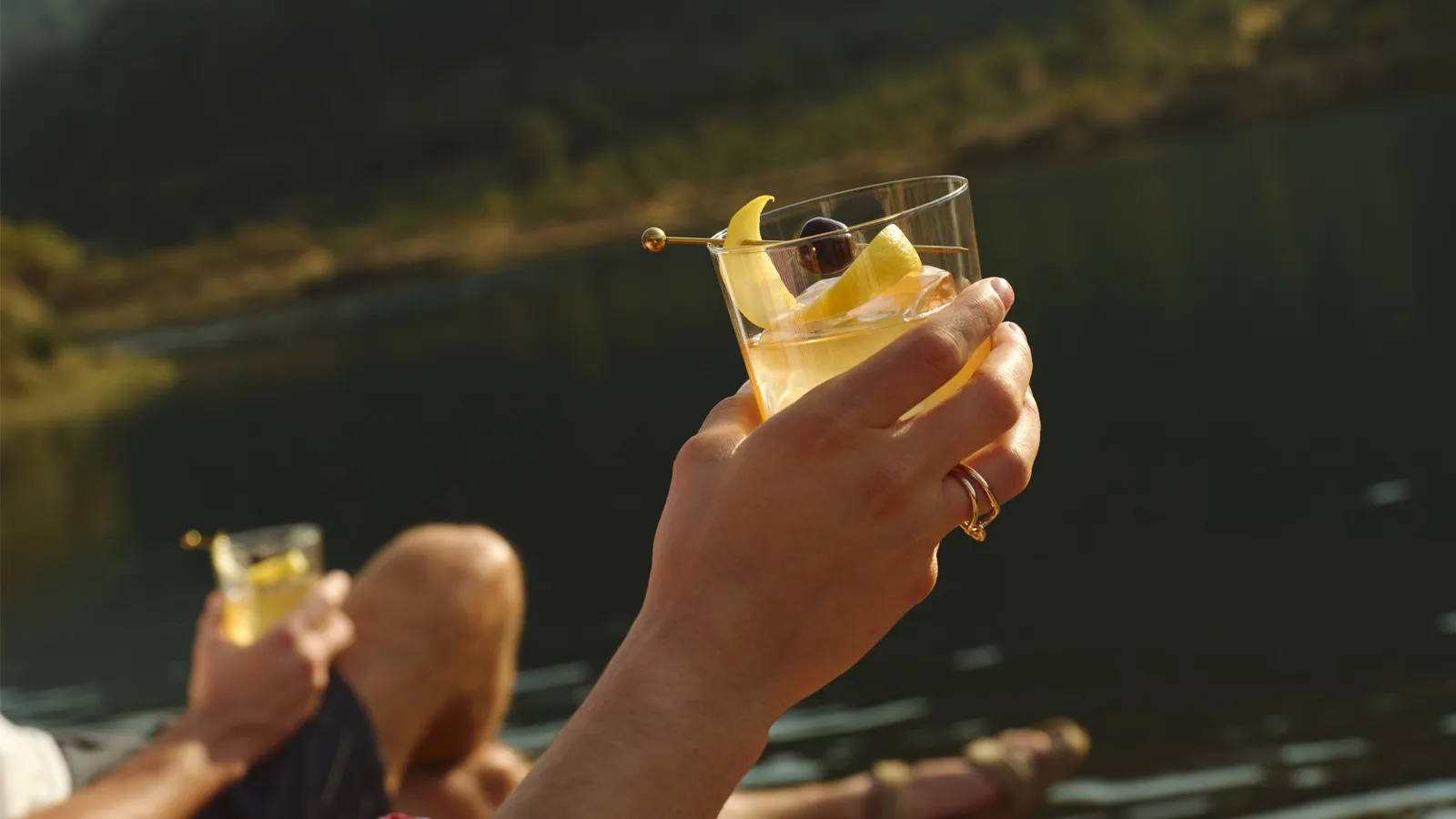 A hand holds a garnished cocktail overlooking a lake, Whisky Maker's Mark.