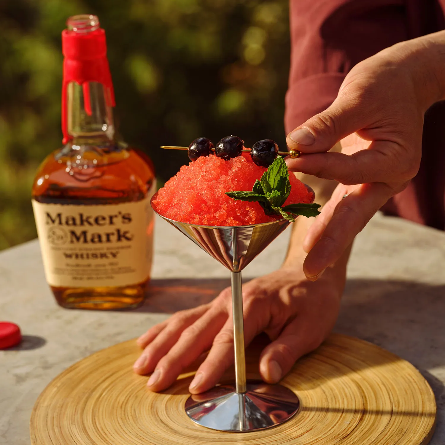 A person garnishes a Whisky Cherry Cone cocktail with cherries and mint, next to a bottle of Whisky Maker's Mark, Cocktail Whisky Maker's Mark.
