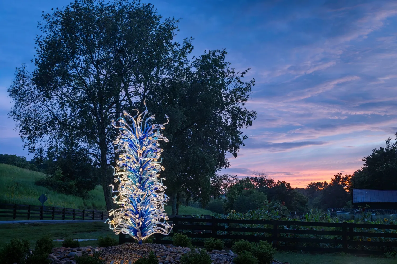 A towering blue and white glass sculpture stands amidst lush greenery under a twilight sky, framed by trees and a wooden fence.