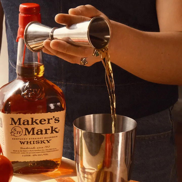 A person pours Maker's Mark bourbon from a jigger into a cocktail shaker, with a tomato and the whiskey bottle in view.