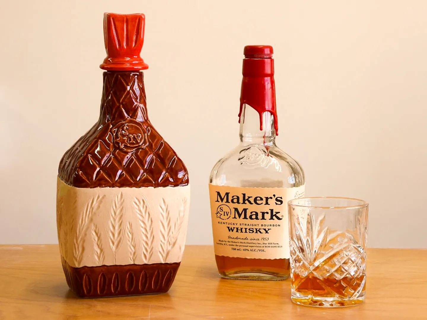 A decorative ceramic decanter next to a bottle of Maker's Mark bourbon and a crystal glass filled with amber whiskey.