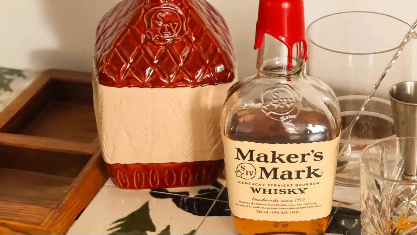 A bottle of Maker's Mark whiskey with a red wax seal beside a decorative ceramic jug, set on a patterned tablecloth with bar tools.