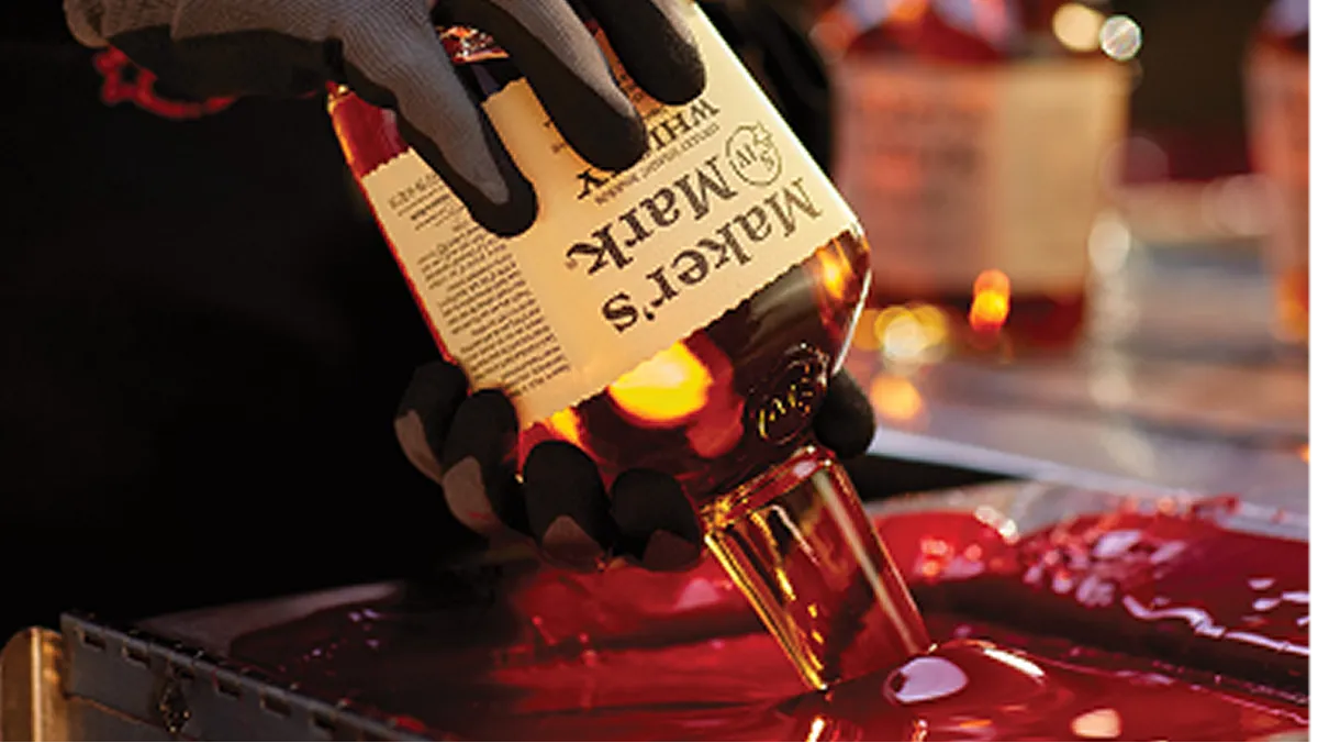 A gloved hand pours whiskey from a Maker's Mark bottle into a pool of red wax, surrounded by blurred whiskey bottles in the background.