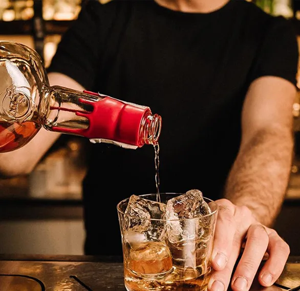 A bartender pours amber liquid from a bottle into a glass filled with ice, set against a softly lit bar background.