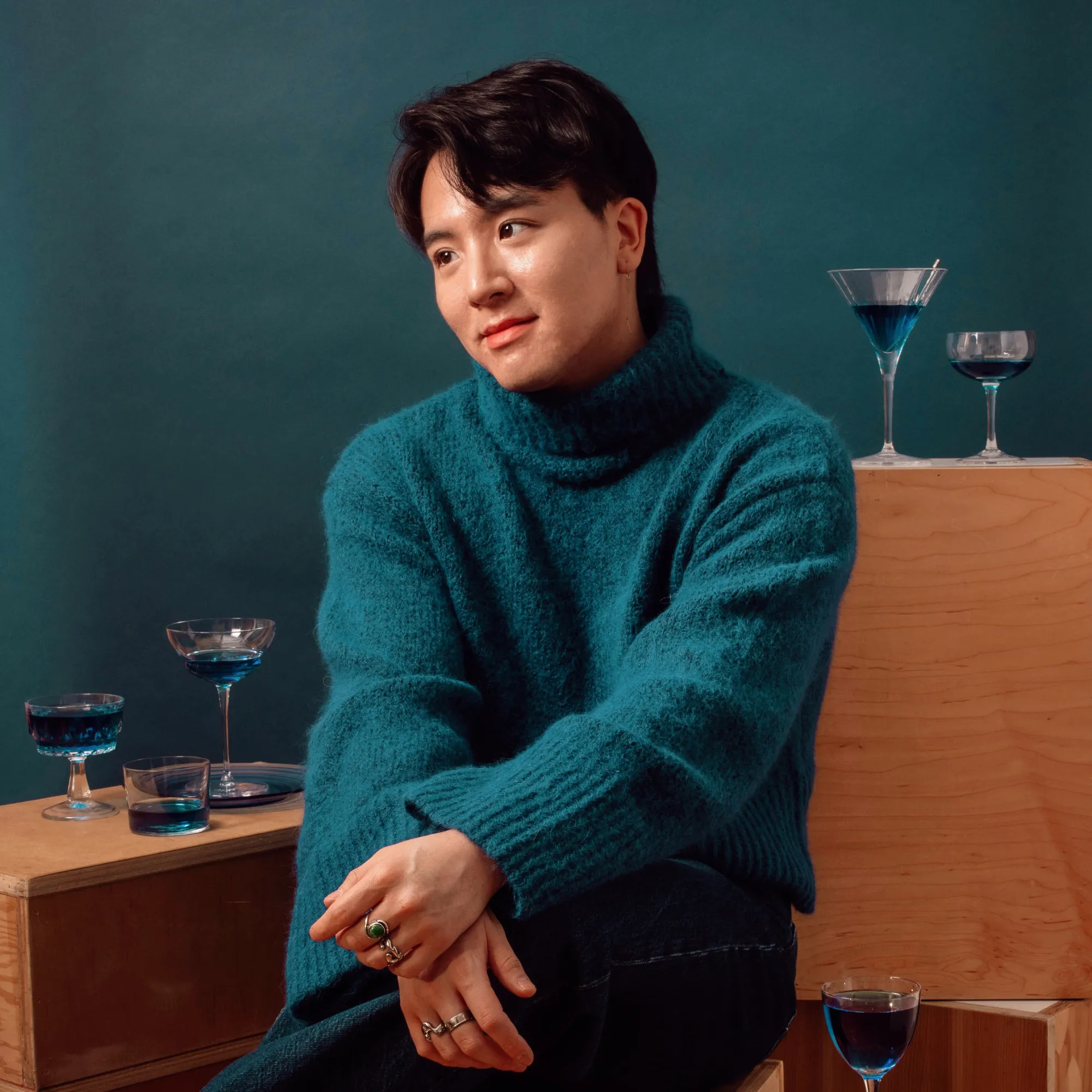 Jae Bae with dark hair styled casually, wearing a cozy teal turtleneck sweater, against a smooth teal background.