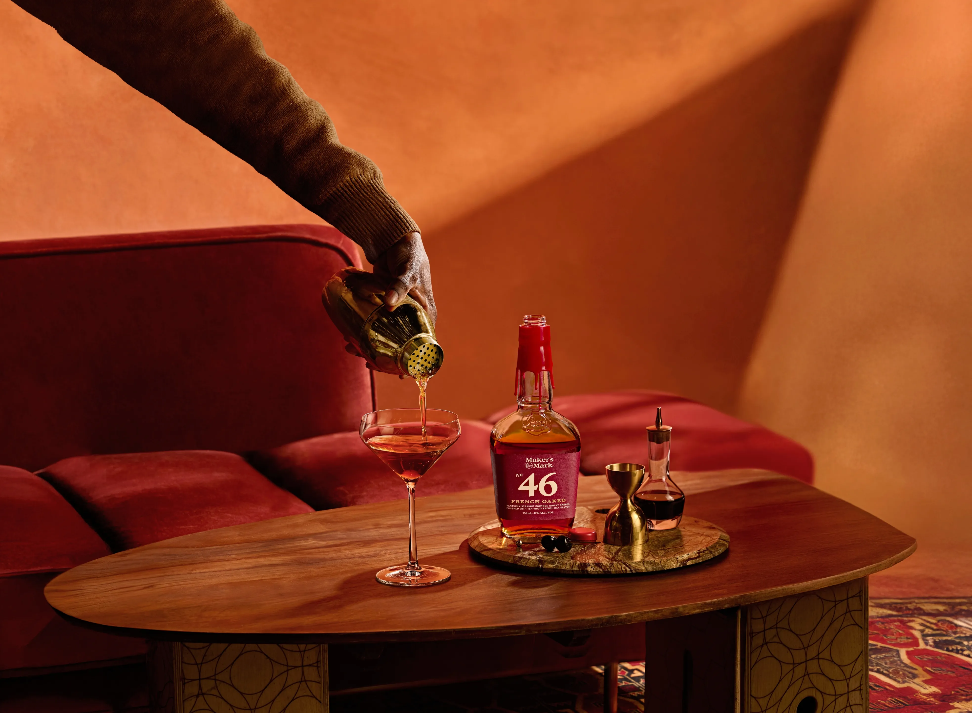 A hand pours whiskey from a shaker into a glass beside a bottle of Maker's Mark 46 on a wooden table, with a cozy red couch in the background.