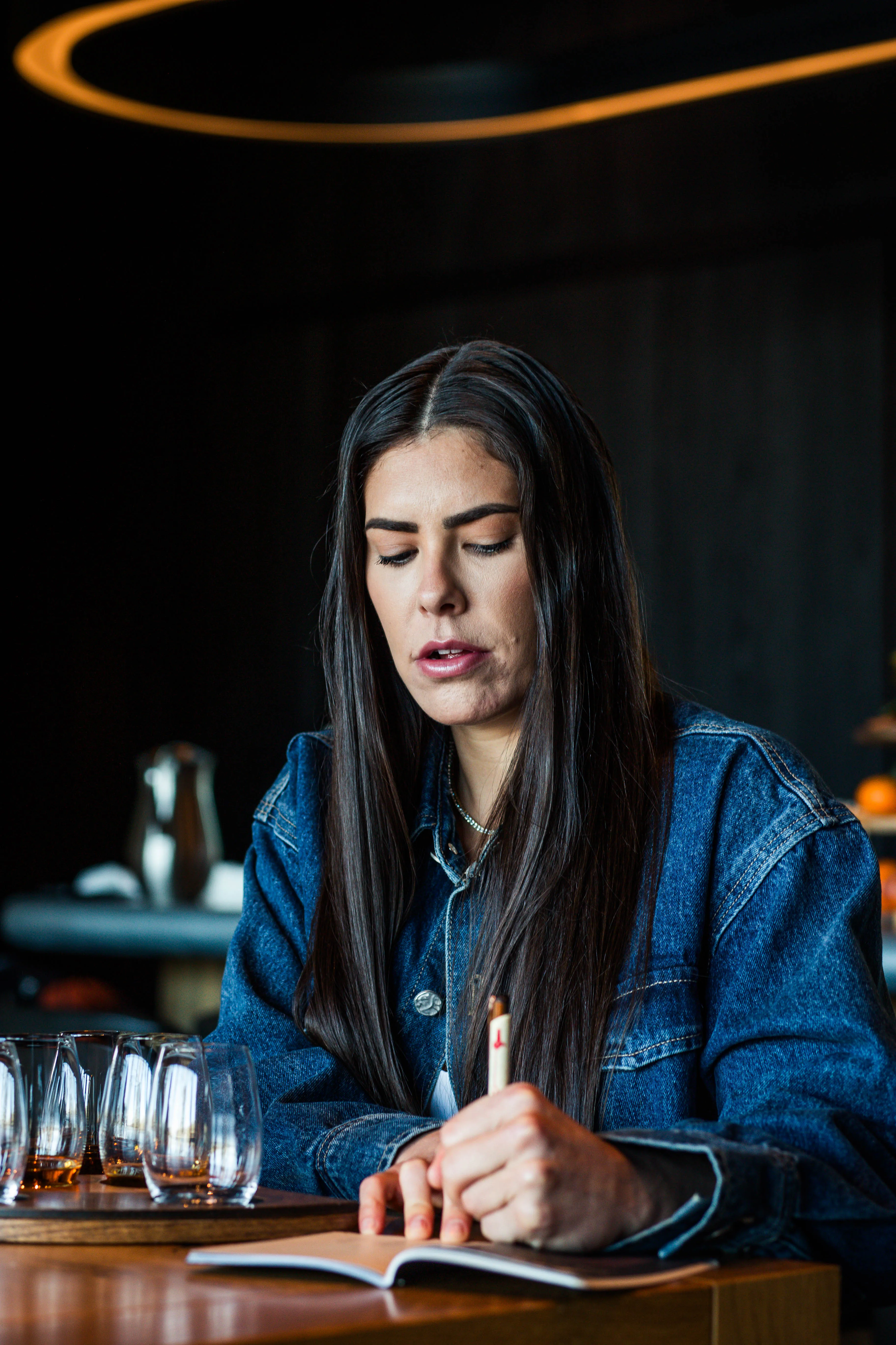 Kelsey Plum wearing safety glasses and a black apron dips a bourbon bottle into a vat of signature red wax, focusing intently as the liquid drips down the neck, Whisky Maker’s Mark.