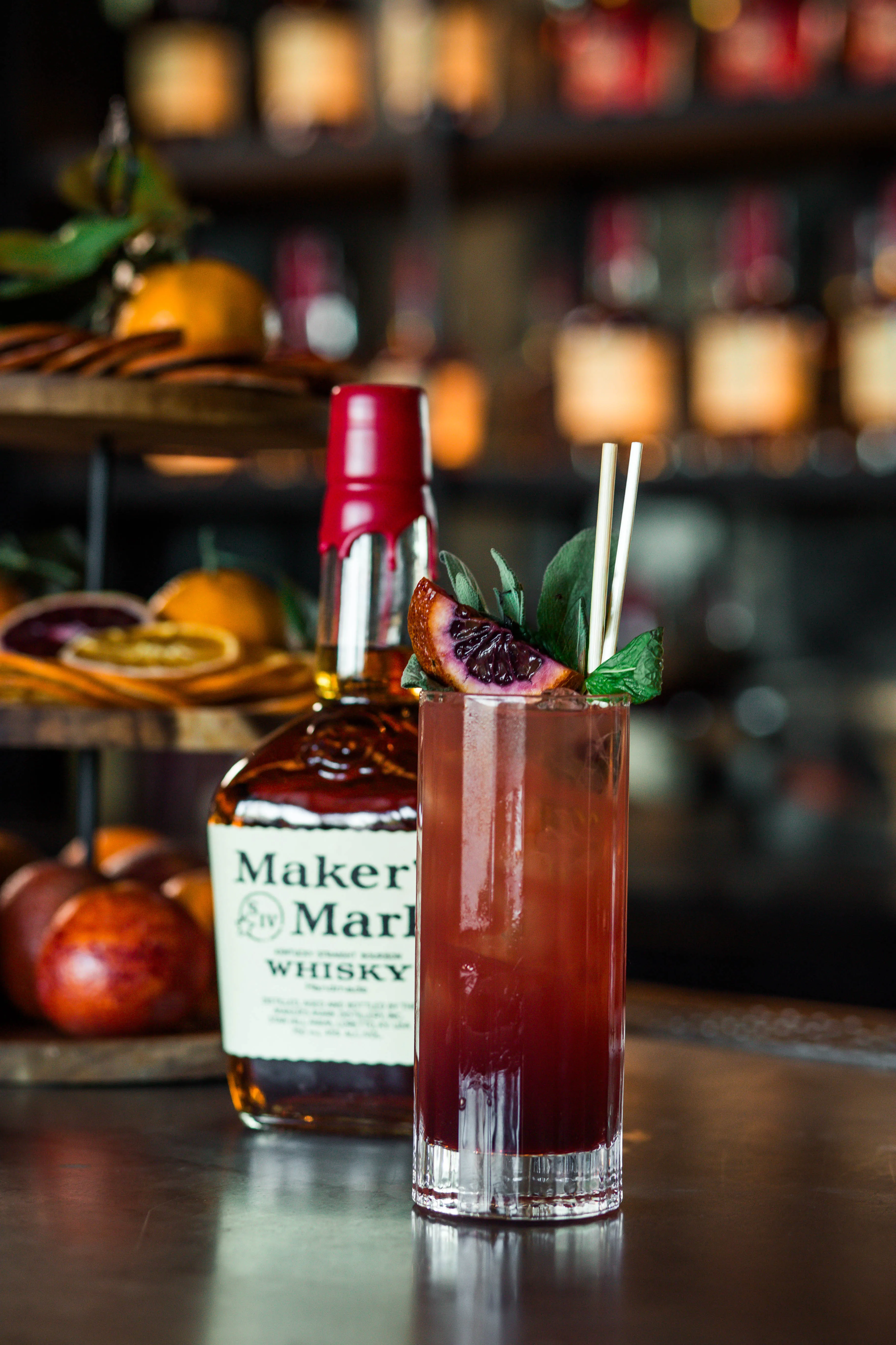 A close-up shot of a classic bourbon bottle with its red wax seal sitting on a bar next to a tall, vibrant red cocktail and a wooden tray of dried citrus wheels and fresh fruit, Whisky Maker’s Mark.