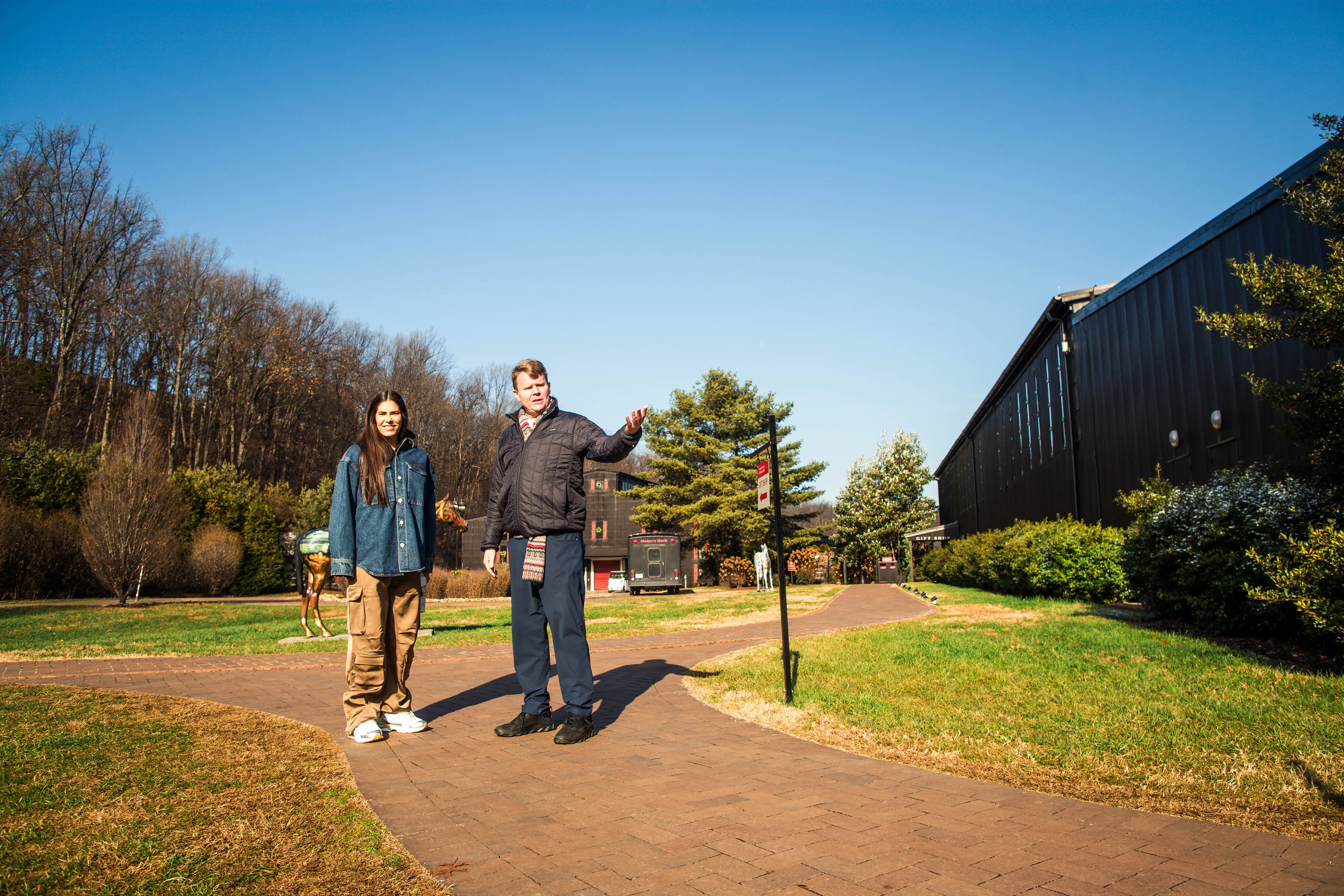 Kelsey Plum and a companion walk along a brick path on the distillery grounds, passing a colorful horse statue under a clear blue sky, Whisky Maker’s Mark.