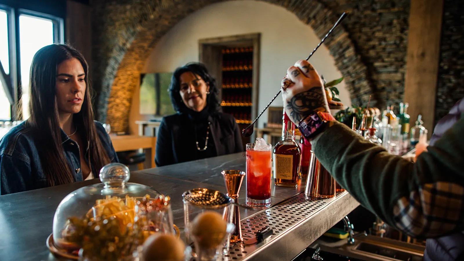 Kelsey Plum and a companion sit at a rustic stone-walled bar, watching a bartender with a tattooed arm expertly craft a red cocktail using a long stirring spoon, Whisky Maker’s Mark.