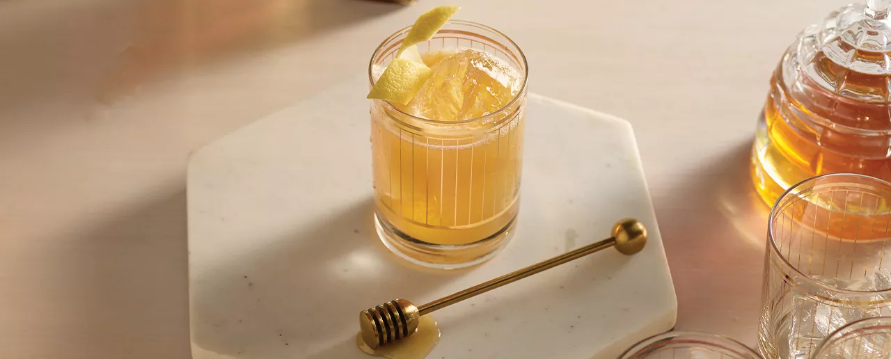 Glass tumbler with a Maker's Mark Gold Rush cocktail, garnished with a lemon peel. On the right are the ingredients.
