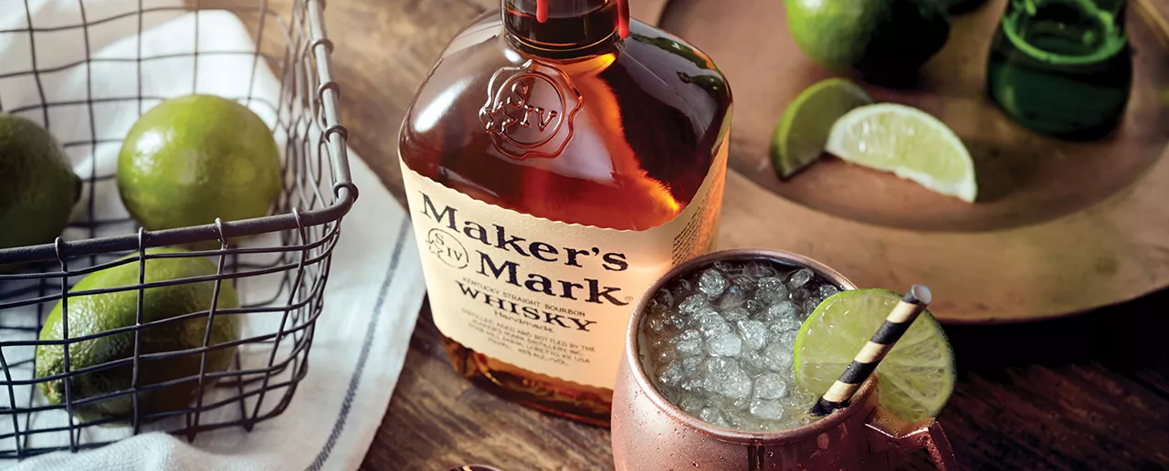 A bottle of Maker's Mark sits next to a copper mug with a Maker's Mule cocktail, garnished with a striped straw and limes. Limes sit on the table behind the bottle. 
