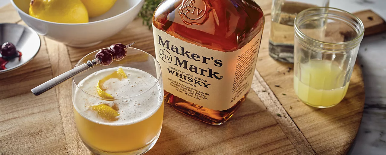 A bottle of Maker's Mark sits on a wooden round tray next to a Whisky Sour in a glass tumbler, garnished with cocktail cherries and lemon peel. Cherries and lemons sit in bowls around the bottle. 