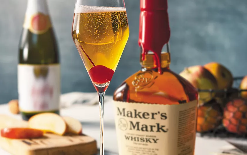 A white wine glass with a Kentucky Bubbly cocktail and garnished with a cherry is next to a handle of Maker's Mark and behind is a bottle of sparkling cider.