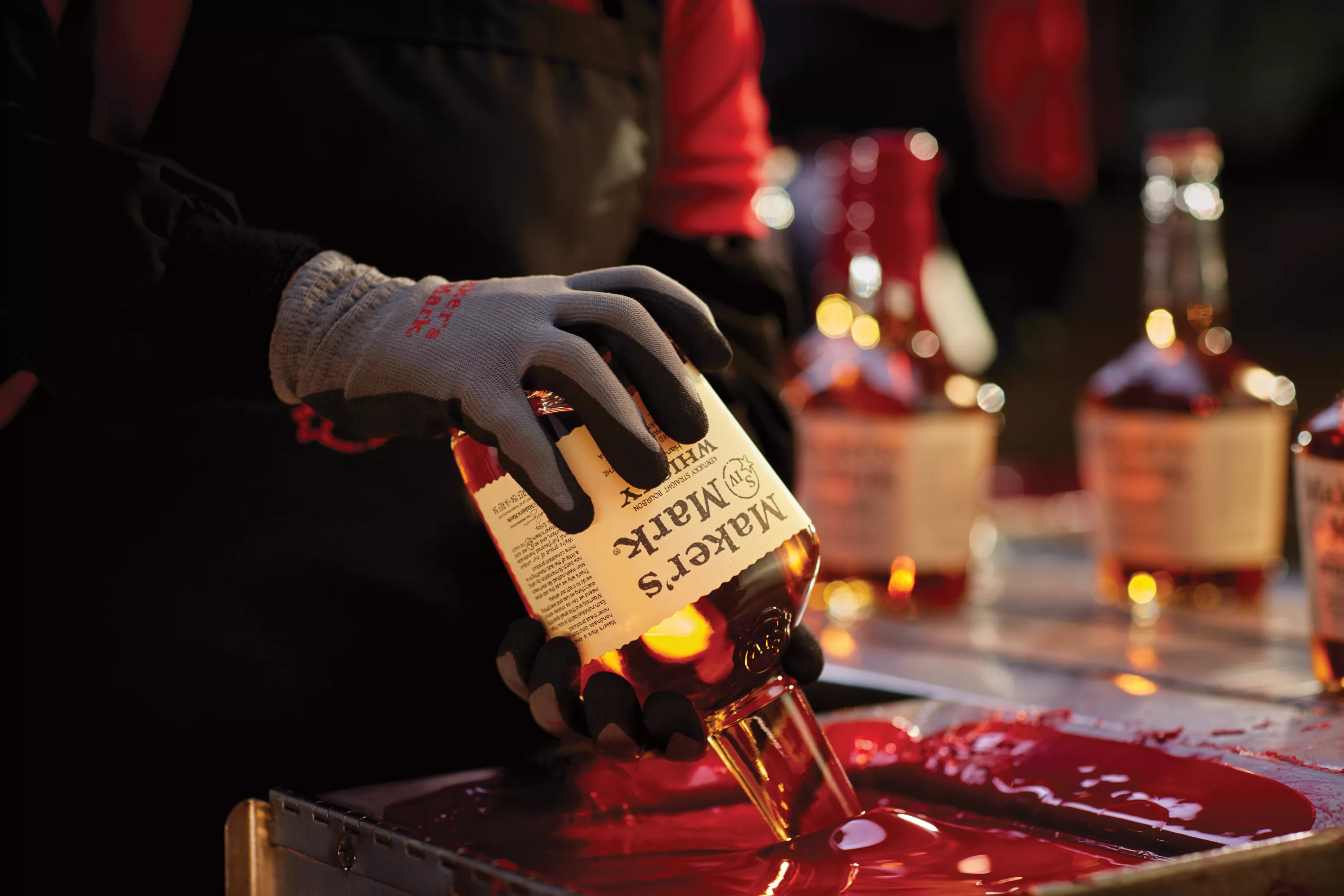 A pair of gloved hands in the left side of the screen are dipping a Maker's Mark bottle, neck down, into a container of hot red wax. There are three bottles of Maker's Mark on a counter to the right.