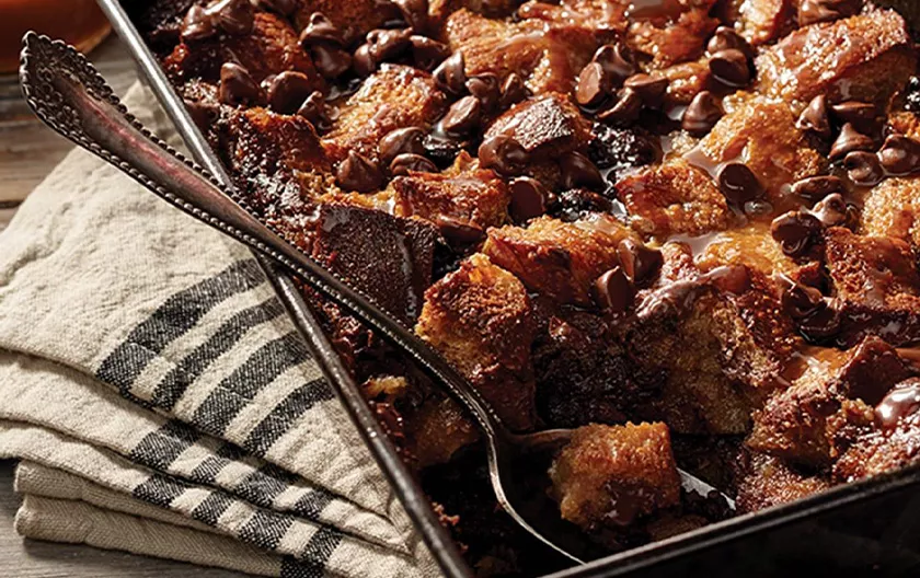 Bread pudding in a square pan with fruit toppings sits next to a bottle of Maker's Mark.