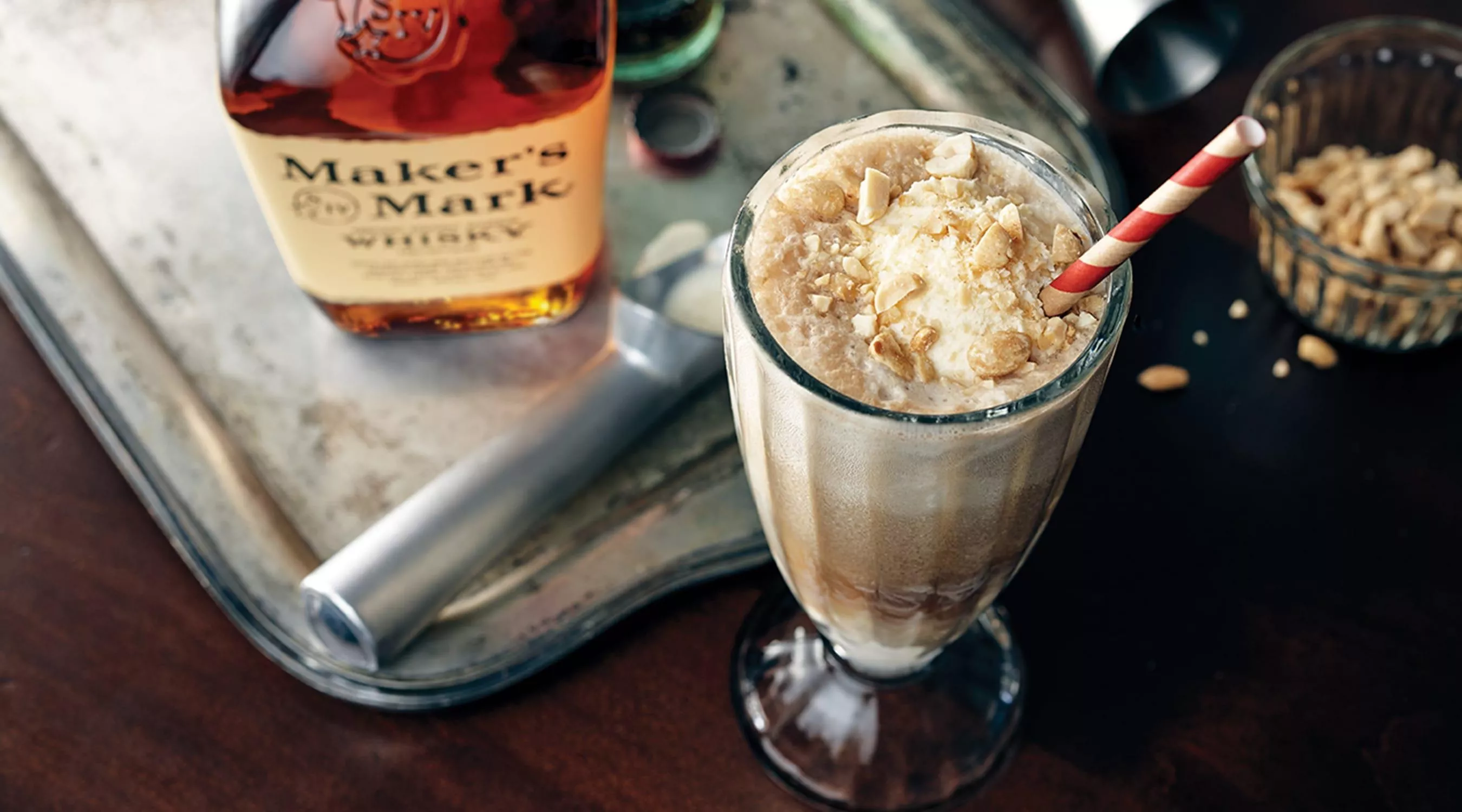 A top down image of a fountain style drink mixed with ice cream and Coca Cola with a red and white straw next to a bottle of classic Maker's Mark. 
