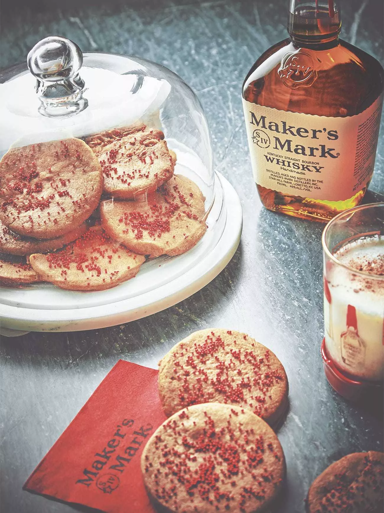 Eggnog spiced butter cookies with red sprinkles displayed in a glass serving platter, with a few cookies on a red napkin next to a bottle of Classic Maker's Mark.