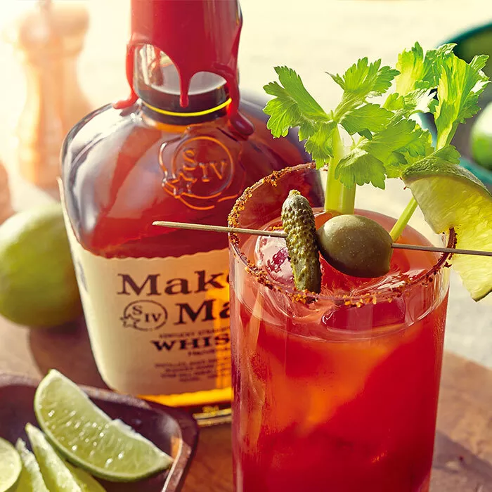 A bottle of Maker's Mark bourbon whisky behind a tall glass with a Bourbon Bloody Mary over ice and a pickle, celery, and lime garnish. A small bowl of lime slices is to the left. 