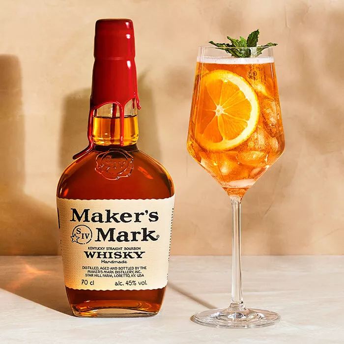 A Maker's Mark Bourbon Spritz cocktail in a wine glass over ice with a lemon garnish and a mint sprig. A Maker's Mark bottle sits to the left of the cocktail. 