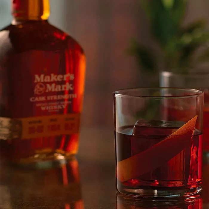 A bottle of Maker's Mark Cask Strength bourbon whisky on the counter next to two tumblers of the Maker's Mark Boulevardier cocktails with orange peel garnishes.