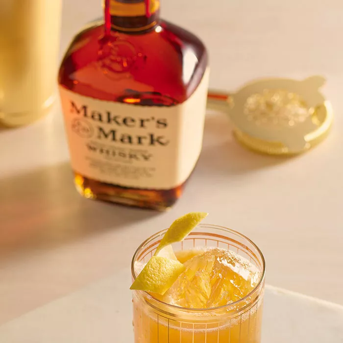 A Maker's Mark Gold Rush cocktail in a tumbler sitting on a large coaster next to a gold honey dipper, glass jar of honey, and three more tumbler glasses.