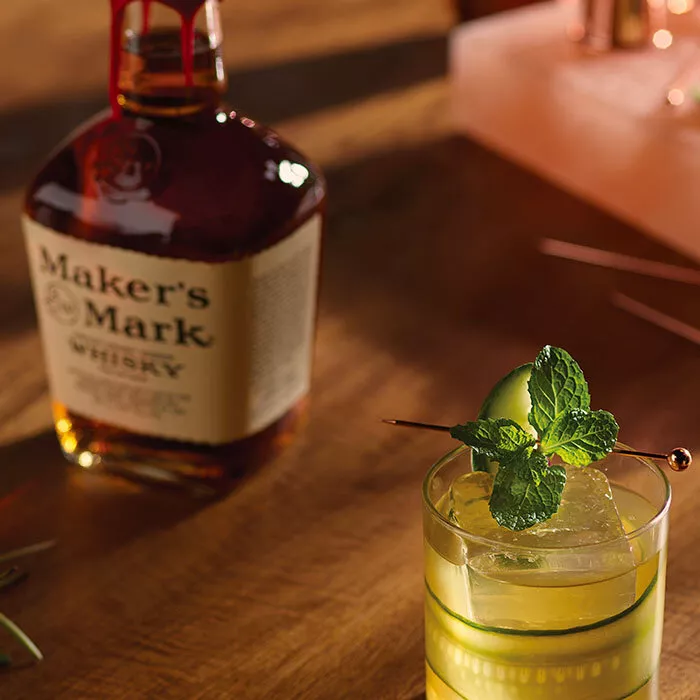 A bottle of Maker's Mark sits next to a Kentucky Made cocktail in a tumbler glass. The cocktail is garnished with cucumber and mint. 