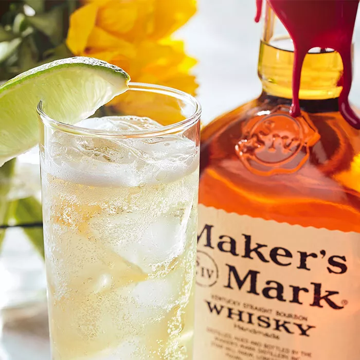 A bottle of Maker's Mark sits behind a tall glass of a Maker's and Ginger cocktail, garnished with a lime. 