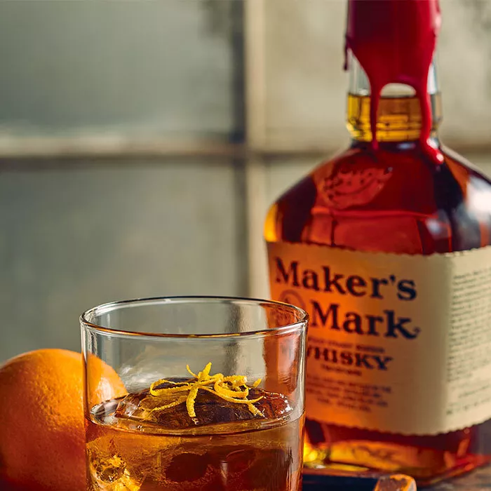 A bottle of Maker's Mark is behind a Maple Old Fashioned in a glass tumbler, garnished with orange peel and cocktail cherries. Pinecones are laid on the table, and an orange sits next to the tumbler.