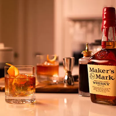 A bottle of Maker's Mark is next to a classic Old Fashioned in a glass tumbler made with Maker's Mark bourbon whisky. The cocktail is garnished with an orange peel.