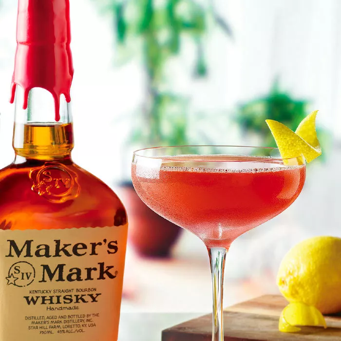 A bottle of Maker's Mark is to the left of a coupe glass with the Maker's Mark Paper Plane cocktail. The cocktail is garnished with a lemon peel and lemons sit behind the cocktail and bottle. 