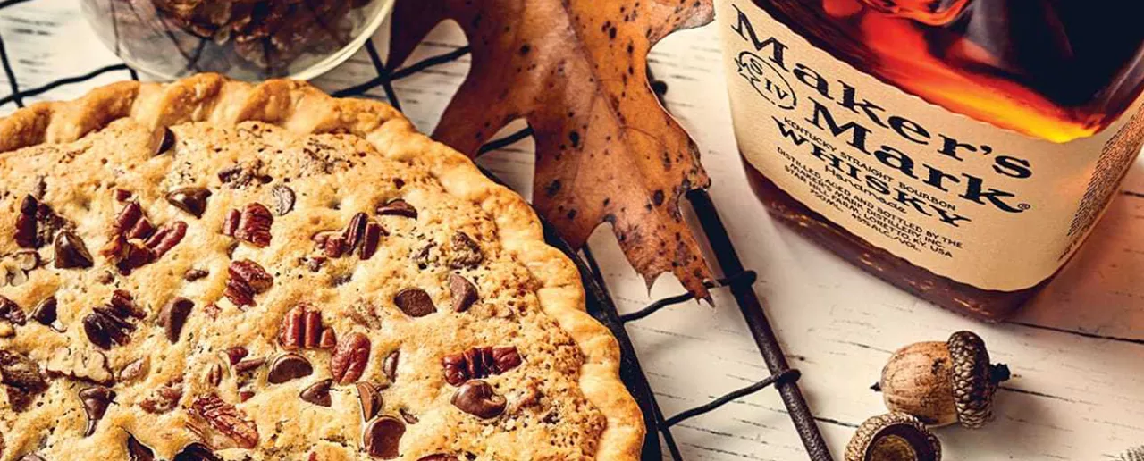 A pecan pie with a slice missing sits on a cooling rack next to a bottle of Maker's Mark Classic.