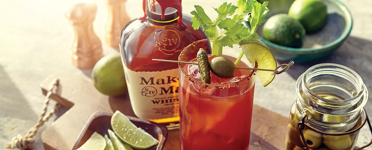 A bottle of Maker's Mark bourbon whisky behind a tall glass with a Bourbon Bloody Mary over ice and a pickle, celery, and lime garnish. A small bowl of lime slices is to the left. 