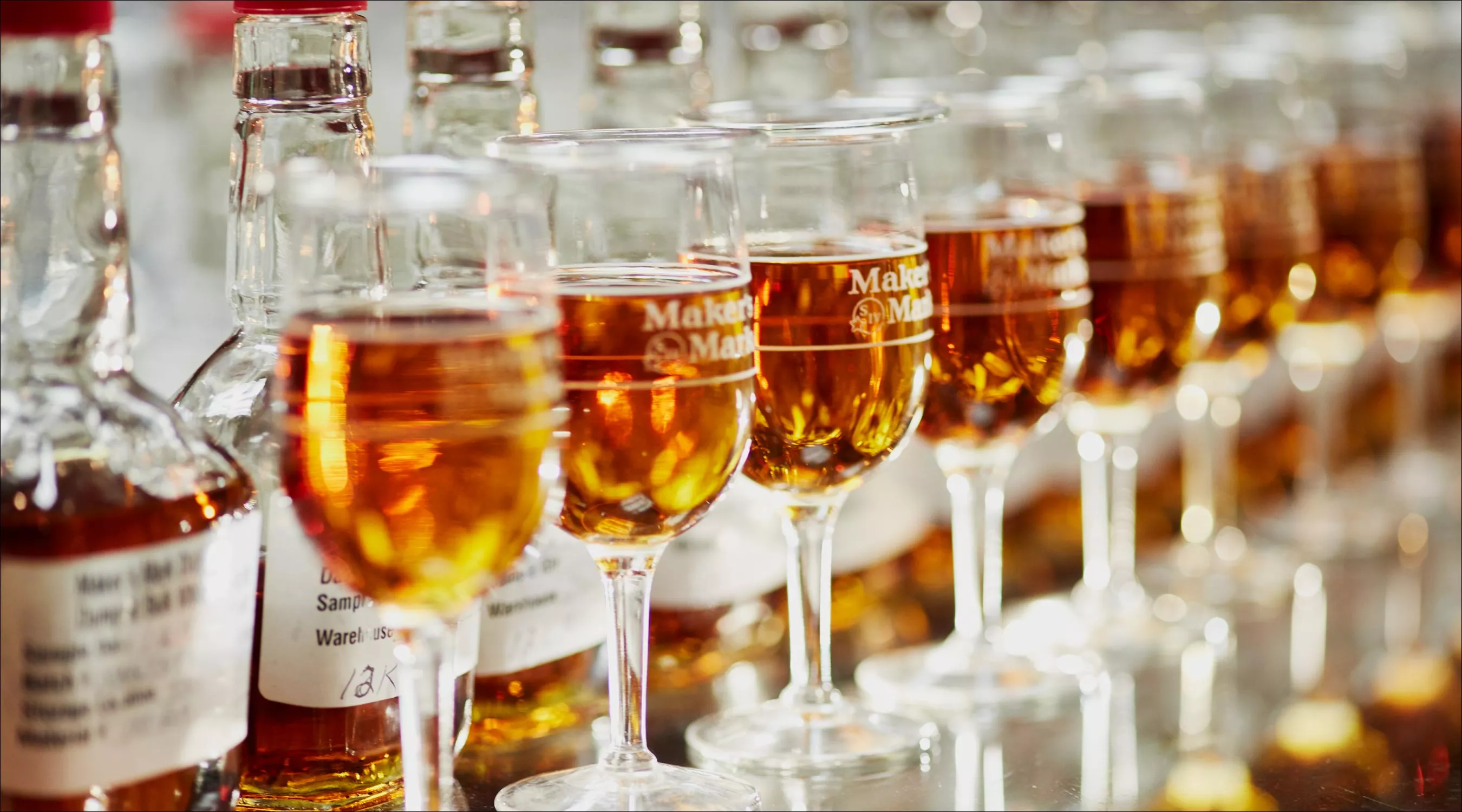 A row of glasses filled with Maker's Mark are presented in front of their bottles.