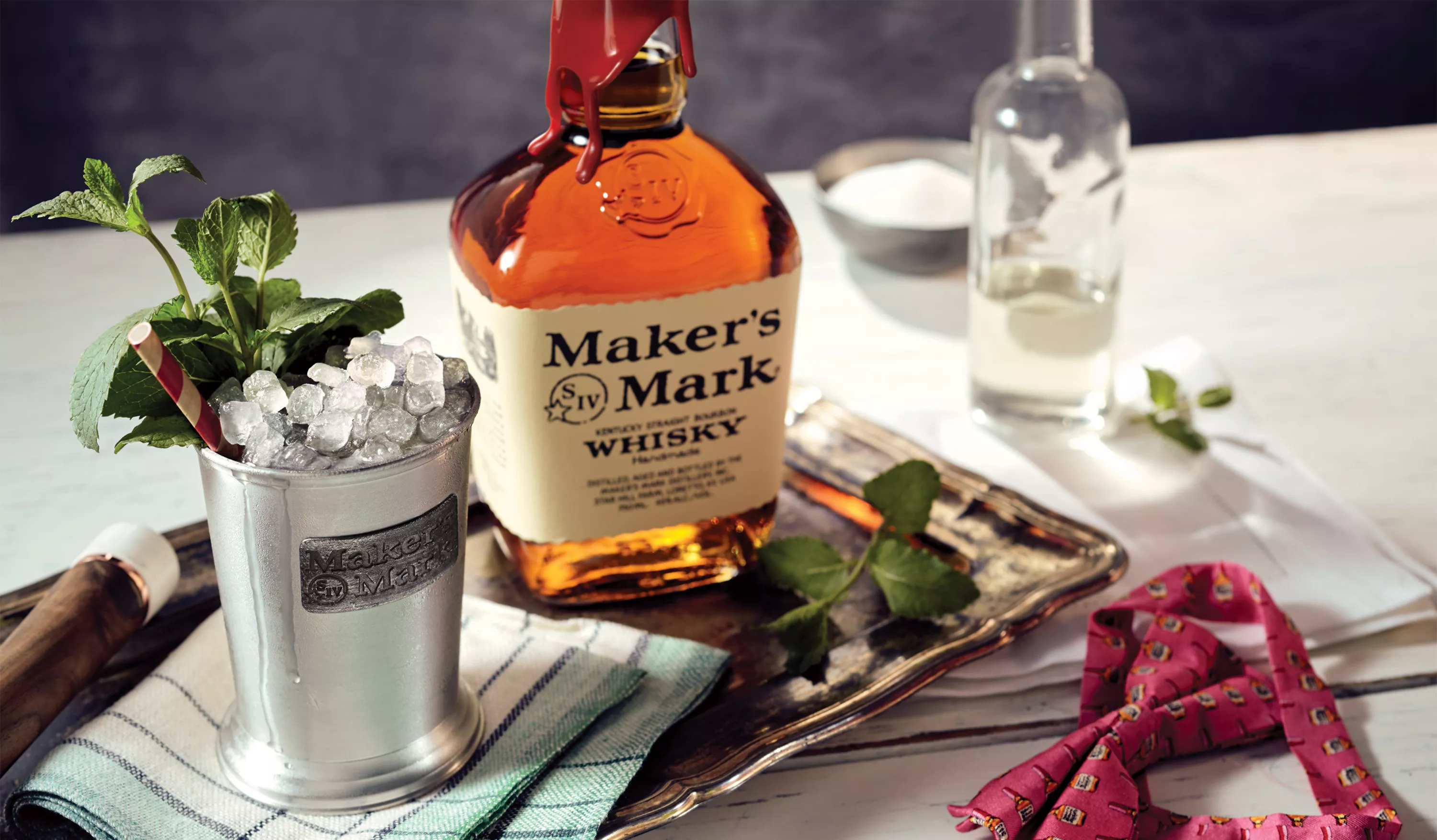 A bottle of Maker's Mark is next to a metal cup with a metal Maker's Mark label on the side. Inside the cup is a Maker's Mark Mint Julep cocktail, garnished with a striped straw and a sprig of mint. 