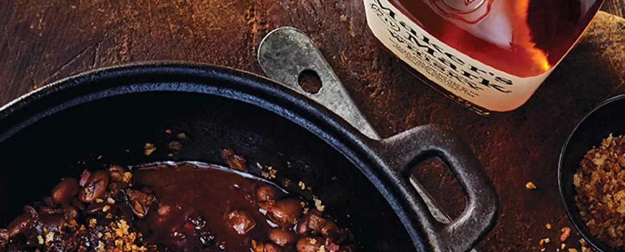A top down image of a cast iron skillet filled with baked beans next to a bottle of Maker's Mark.
