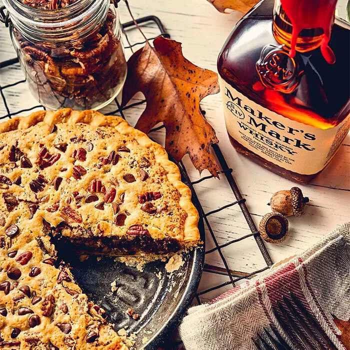 bourbon pecan pie on cooking sheet with jar of pecans next to it on a table also with a bottle of makers mark bourbon