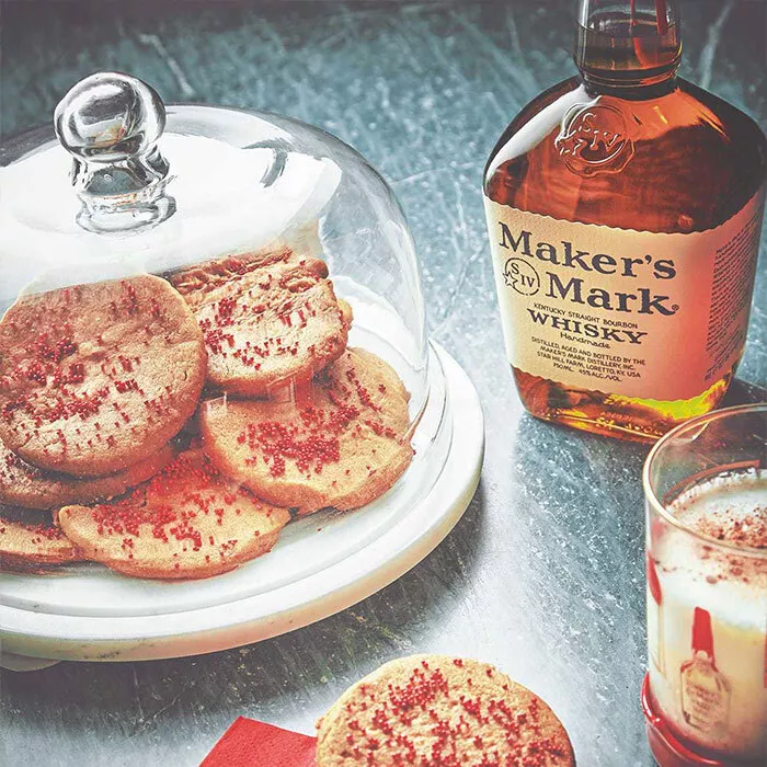 eggnog spiced butter cookies in baking tray covered with glass topper next to bottle of makers mark bourbon