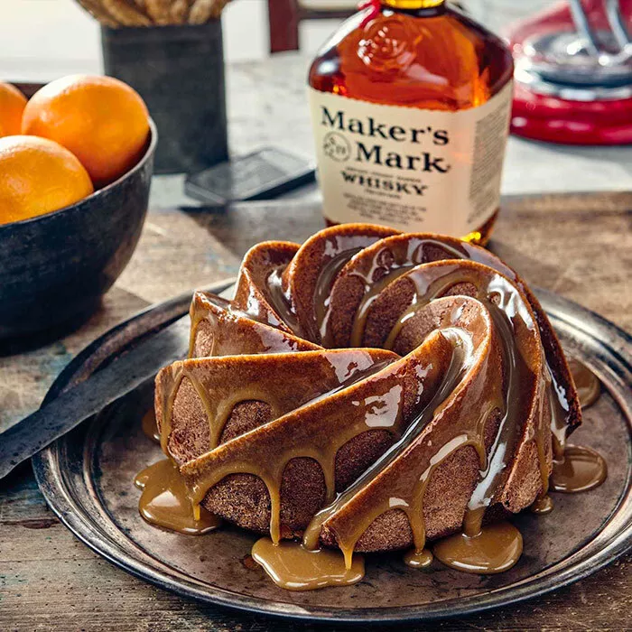 old fashioned bundt cake on plat istting on a table next to bowl of oranges and a bottle of makers a makers mark bourbon